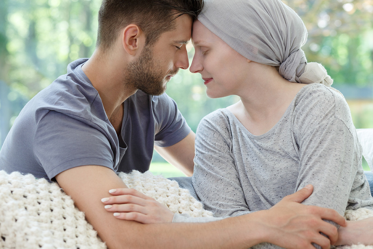 Ein junger Mann und eine Frau mit einem Kopftuch sitzen eng beieinander und lehnen ihre Stirnen sanft aneinander. Beide haben die Augen geschlossen und zeigen einen emotionalen Moment der Nähe und Unterstützung. Die Frau wirkt krankheitsbedingt geschwächt, was auf eine mögliche Krebserkrankung hindeuten könnte. Der Mann hält ihre Arme fürsorglich, was Trost und Verbundenheit ausdrückt.