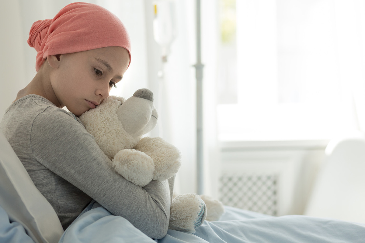 Nachdenkliches Kind mit rosa Kopftuch sitzt in einem Krankenhausbett und hält einen weißen Plüschbären fest umarmt. Im Hintergrund ist ein Infusionsständer zu sehen, der auf eine medizinische Behandlung hinweist. Das Kind trägt ein graues Langarmshirt und blickt traurig nach unten. Die Szene vermittelt eine ruhige, nachdenkliche Stimmung.