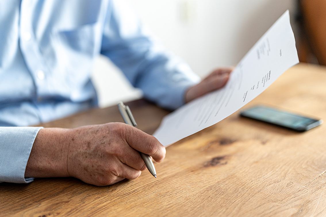 Nahaufnahme einer Hand, die ein Blatt Papier hält, während die andere Hand einen Stift umfasst. Die Person trägt ein hellblaues Hemd und sitzt an einem Holztisch. Im Hintergrund liegt ein Smartphone unscharf auf dem Tisch.