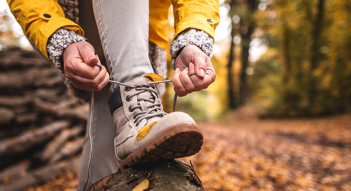Eine Person bindet die Schnürsenkel ihrer beigefarbenen Wanderschuhe, während sie den Fuß auf einen Baumstamm im herbstlichen Wald stellt. Die Person trägt eine gelbe Jacke mit gestrickten grauen Ärmeln. Auf den Schuhen und am Boden liegen bunte Herbstblätter, der Hintergrund zeigt unscharfe Bäume in warmen Gelb- und Brauntönen.