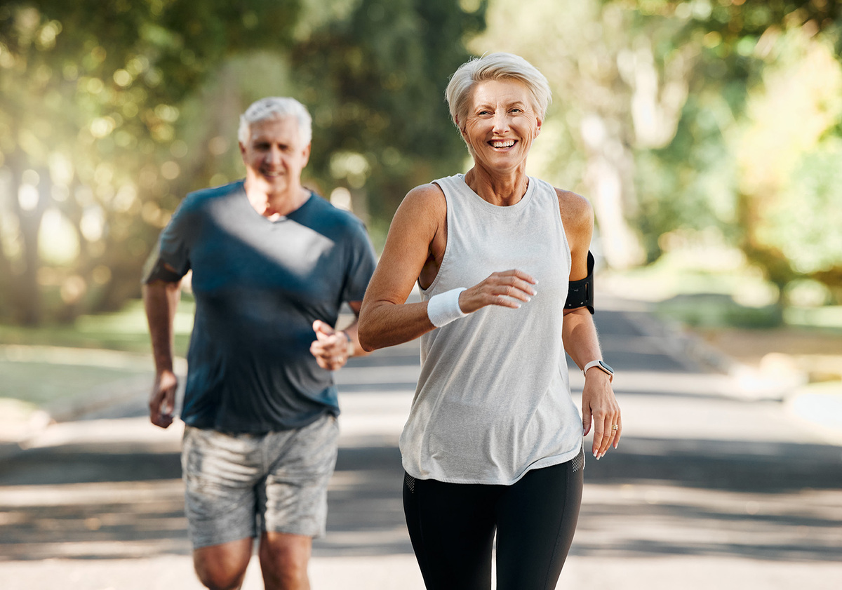 Ein älteres, sportlich aktives Paar joggt draußen auf einer von Bäumen gesäumten Straße an einem sonnigen Tag. Die Frau im Vordergrund trägt ein ärmelloses, helles Oberteil, eine schwarze Leggings und ein Fitness-Armband am Oberarm. Sie lächelt, während sie läuft. Der Mann im Hintergrund trägt ein dunkles T-Shirt und graue Shorts. Beide wirken motiviert und fit.