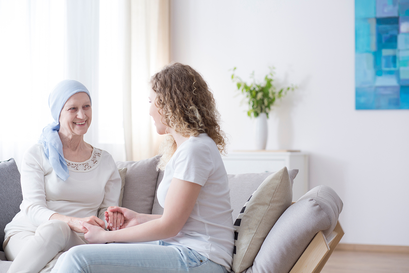 Junge Frau sitzt auf einem Sofa und hält die Hände einer älteren Frau mit einem hellblauen Kopftuch. Beide lächeln sich warm an. Die Szene spielt in einem hellen, freundlich eingerichteten Raum mit Pflanzen im Hintergrund.