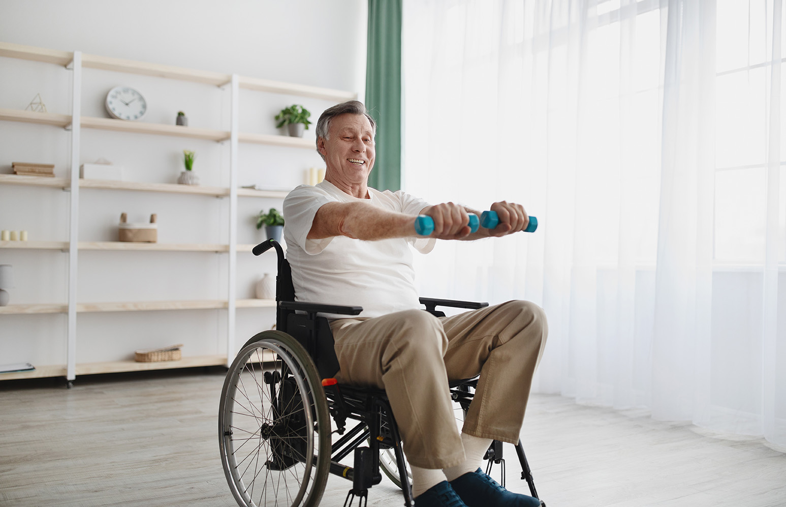 Älterer Mann im Rollstuhl trainiert mit kleinen Hanteln in einem hellen, modern eingerichteten Raum. Er lächelt und scheint motiviert, seine körperliche Fitness zu stärken.