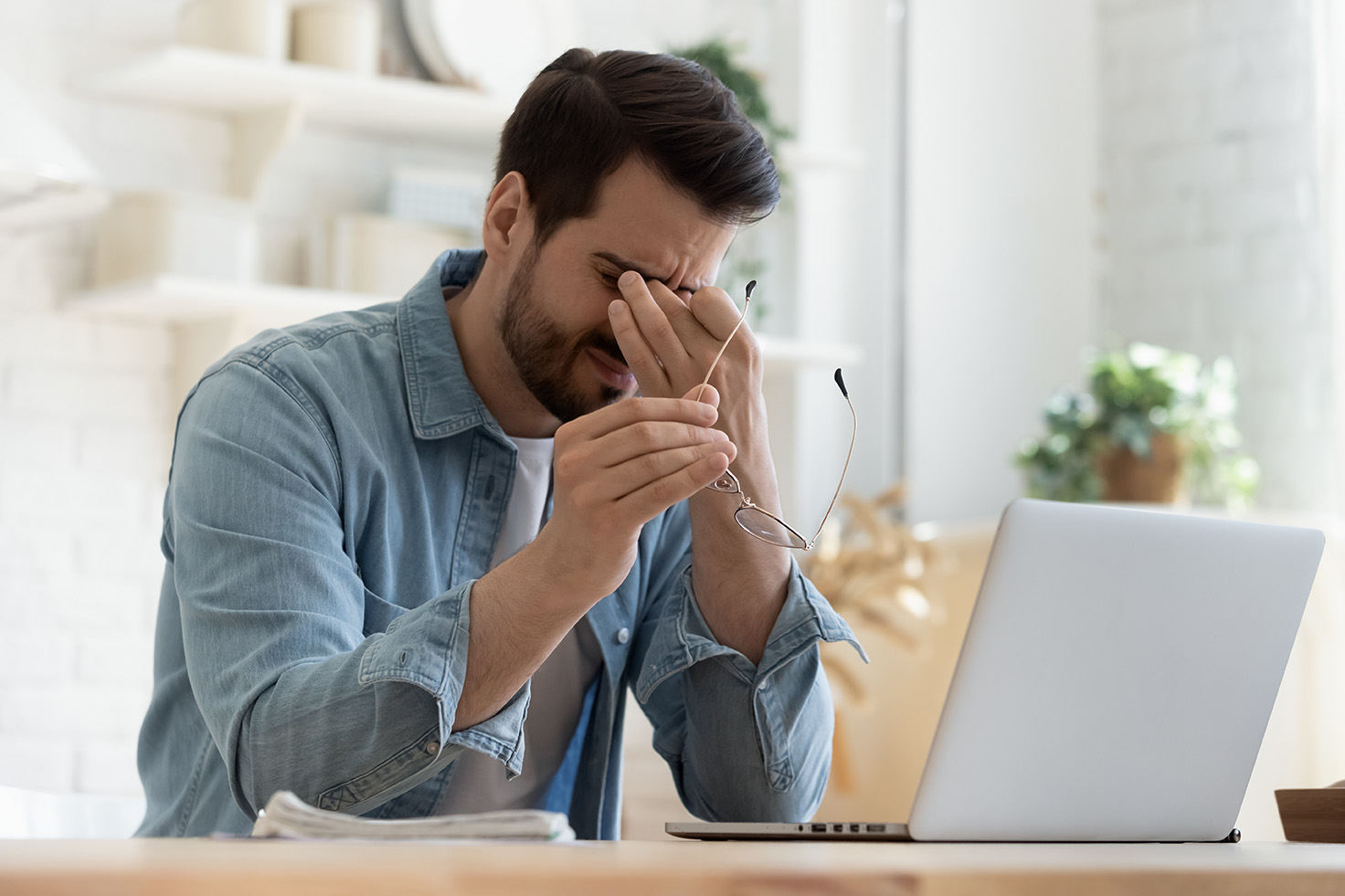 Ein junger Mann sitzt vor einem Laptop, hält seine Brille in der Hand und reibt sich erschöpft die Augen. Er wirkt gestresst oder müde, möglicherweise durch lange Bildschirmarbeit oder Überanstrengung der Augen.