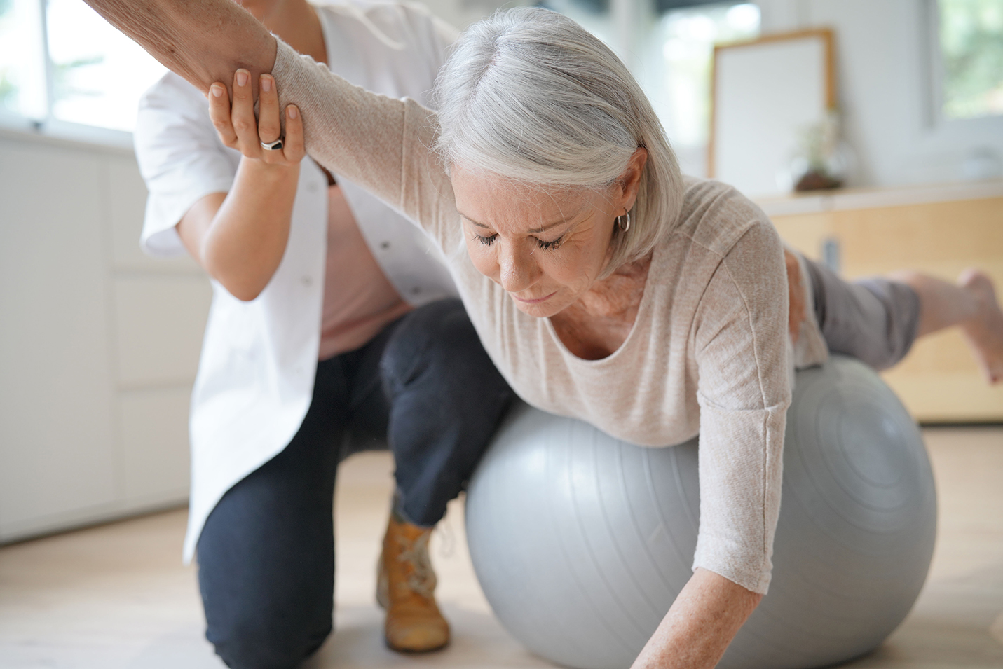 Ältere Frau mit grauen Haaren führt eine Physiotherapie-Übung auf einem Gymnastikball durch. Sie liegt mit dem Bauch auf dem Ball und streckt einen Arm nach vorne, während eine Therapeutin sie unterstützt und ihren Arm stabilisiert. Die Szene spielt in einem hellen Raum mit modernen Möbeln.