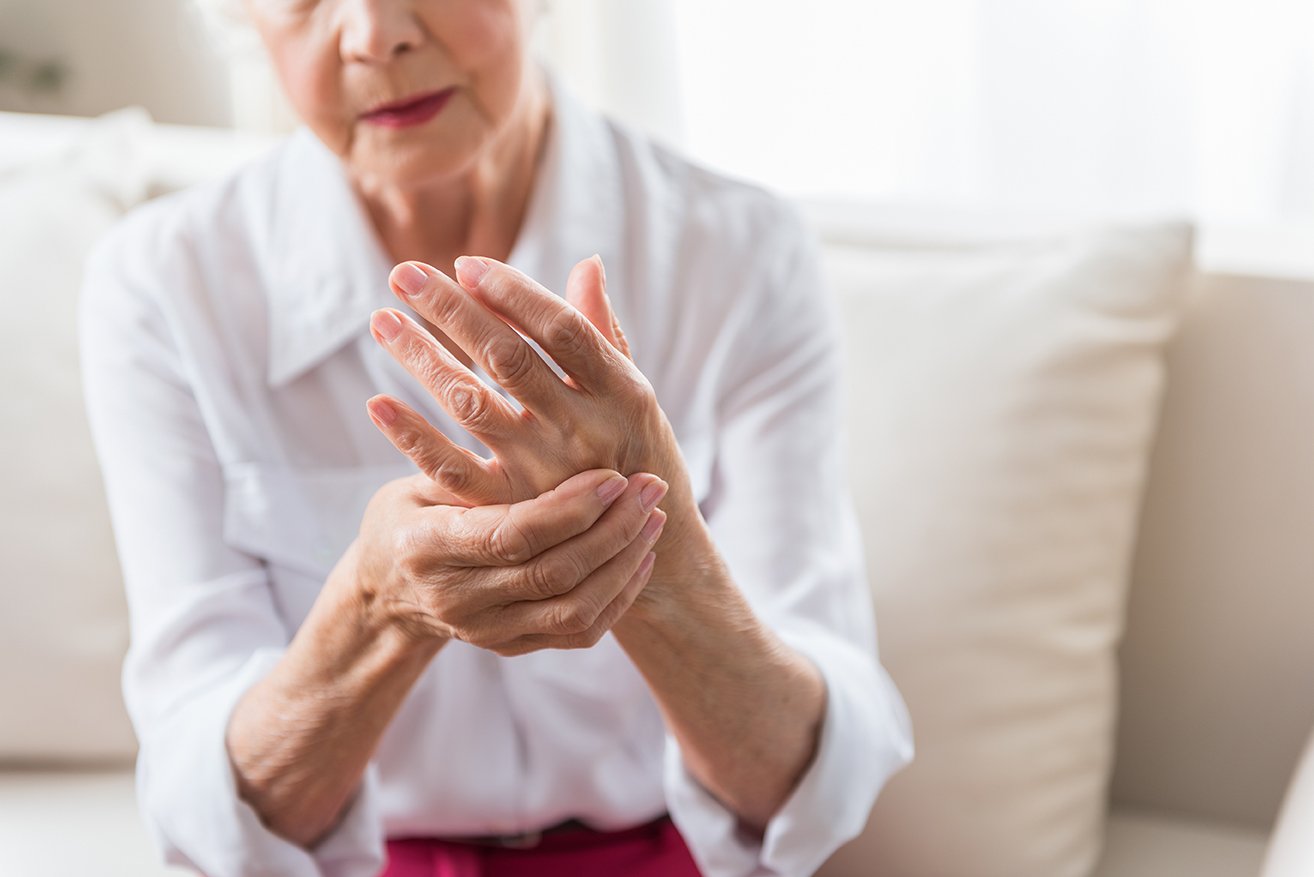 Ältere Frau in weißer Bluse sitzt auf einem Sofa und hält sich mit schmerzverzerrtem Gesichtsausdruck die schmerzende Hand. Sie massiert ihre Finger, was auf Beschwerden wie Arthritis oder Gelenkschmerzen hindeutet. Im Hintergrund sind helle Kissen und ein weiches Sofa zu sehen.