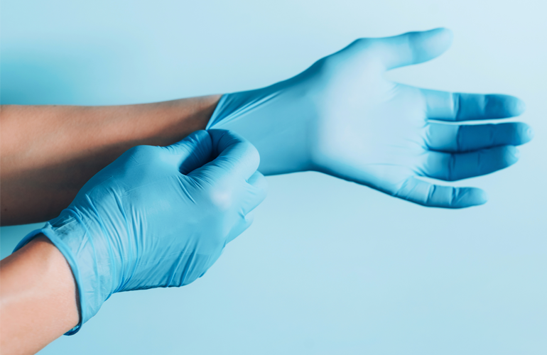 Nahaufnahme von Händen, die blaue Einweghandschuhe anziehen. Eine Hand zieht den Handschuh über das Handgelenk der anderen Hand, um einen sicheren Sitz zu gewährleisten. Der Hintergrund ist in einem hellblauen Farbton gehalten.