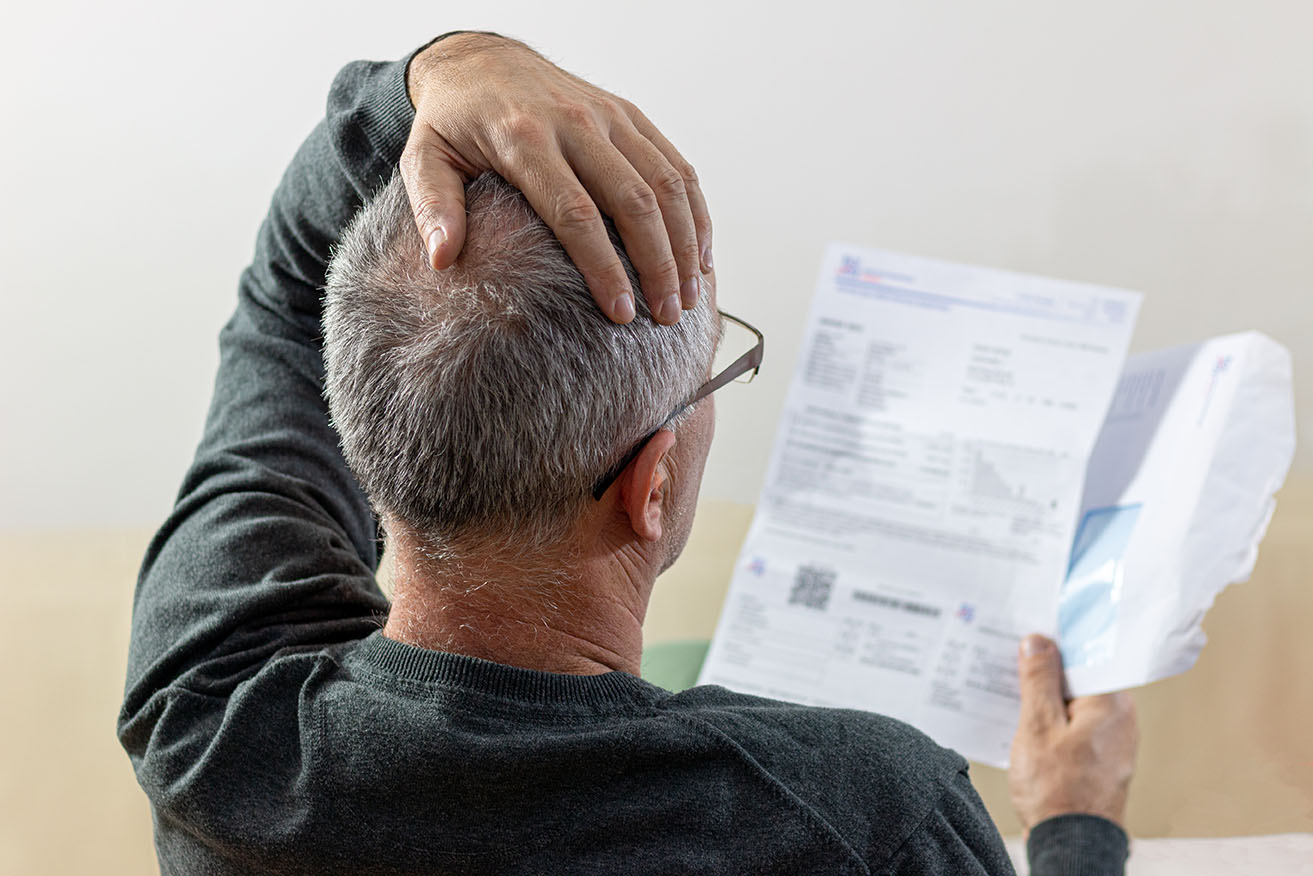 Ein älterer Mann mit grauem Haar und Brille sitzt mit dem Rücken zur Kamera und hält sich besorgt den Kopf, während er einen offiziellen Brief in der Hand hält. Neben ihm liegt ein geöffneter Umschlag. Das Bild vermittelt Sorgen oder Unsicherheit, möglicherweise über eine Rechnung oder einen behördlichen Bescheid.
