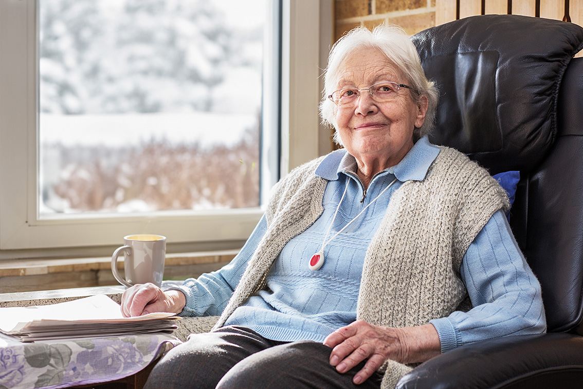 Lächelnde ältere Frau mit weißem Haar und Brille sitzt in einem gemütlichen Sessel vor einem Fenster mit winterlicher Schneelandschaft. Sie trägt eine hellblaue Strickjacke, eine beige Weste und ein Hausnotrufsystem um den Hals. Neben ihr auf einem Tisch liegen Zeitungen und eine Tasse Tee.