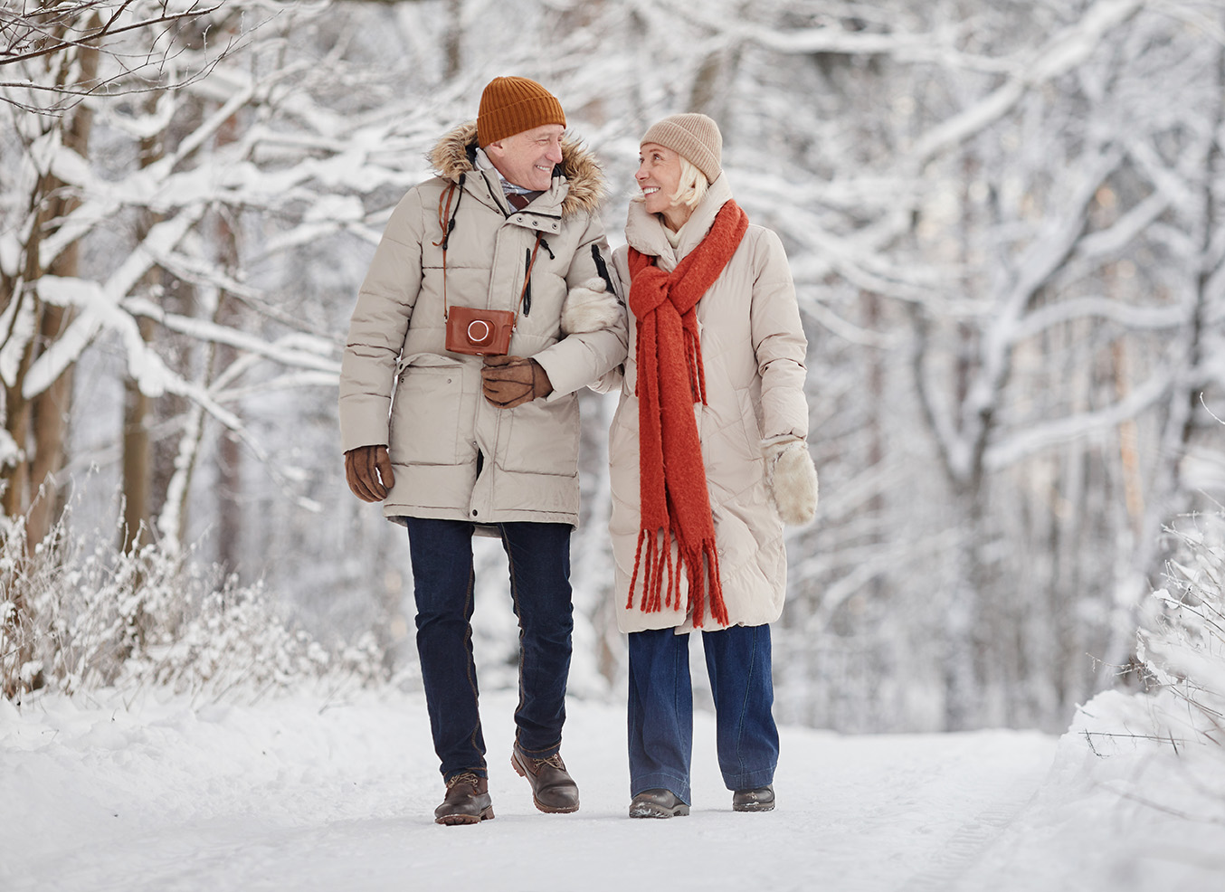 Ein älteres Paar spaziert lächelnd Arm in Arm durch eine verschneite Winterlandschaft. Beide tragen warme Winterkleidung mit Mützen, Schals und dicken Jacken. Der Mann hat eine braune Umhängetasche, die Frau einen auffällig roten Schal. Im Hintergrund sind schneebedeckte Bäume zu sehen, die eine idyllische, winterliche Atmosphäre schaffen.