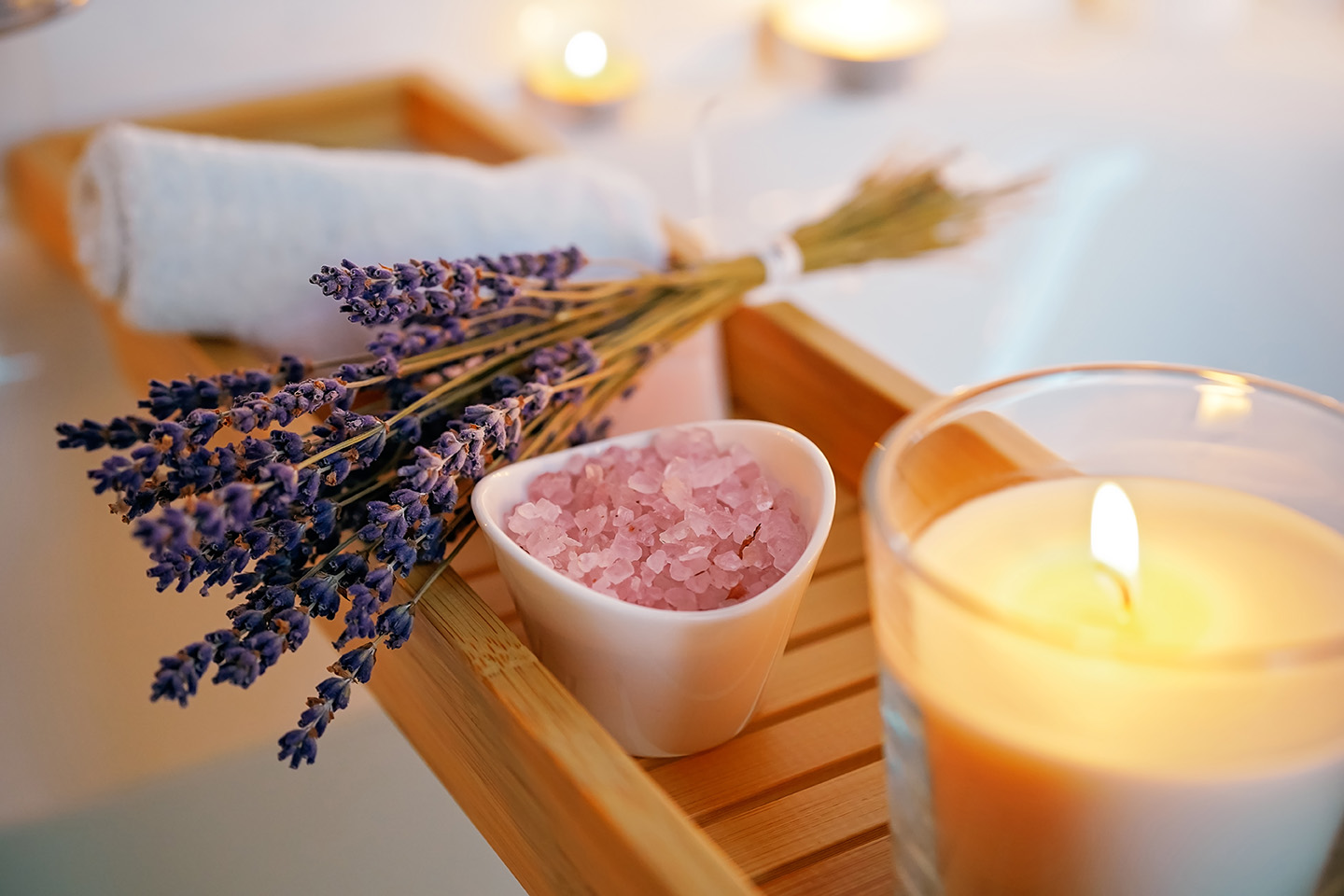Entspannende Wellness-Szene mit einem Bündel getrocknetem Lavendel, einer Schale mit rosa Badesalz und einer brennenden Duftkerze auf einer hölzernen Badewannenablage. Im Hintergrund sind ein gerolltes Handtuch und Kerzenlicht zu sehen.