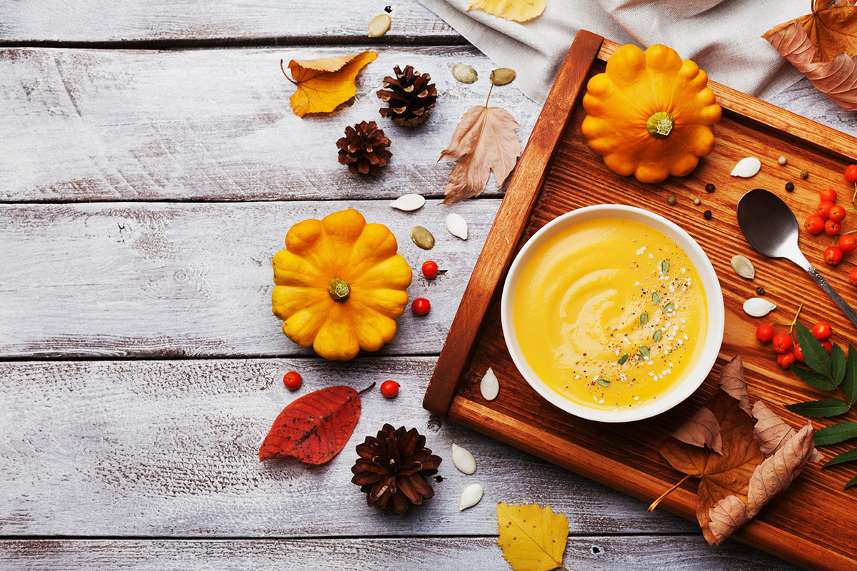 Herbstliche Szene mit einer Schale cremiger Kürbissuppe, garniert mit Kräutern und Samen, auf einem rustikalen Holztablett. Umgeben von kleinen Zierkürbissen, bunten Herbstblättern, Tannenzapfen, Beeren und Kürbiskernen, die auf einer weiß gestrichenen Holzoberfläche verstreut sind.