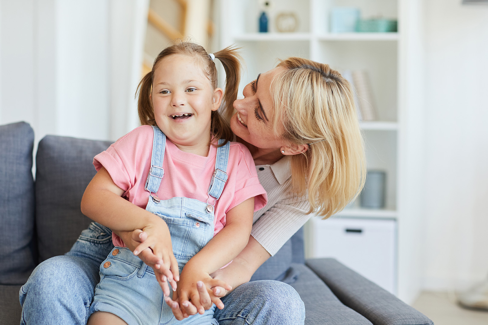 Fröhliches junges Mädchen mit Down-Syndrom lacht, während es auf dem Schoß einer liebevollen Frau sitzt, die sie umarmt. Die Szene spielt sich in einem gemütlichen Wohnzimmer mit heller, freundlicher Atmosphäre ab.