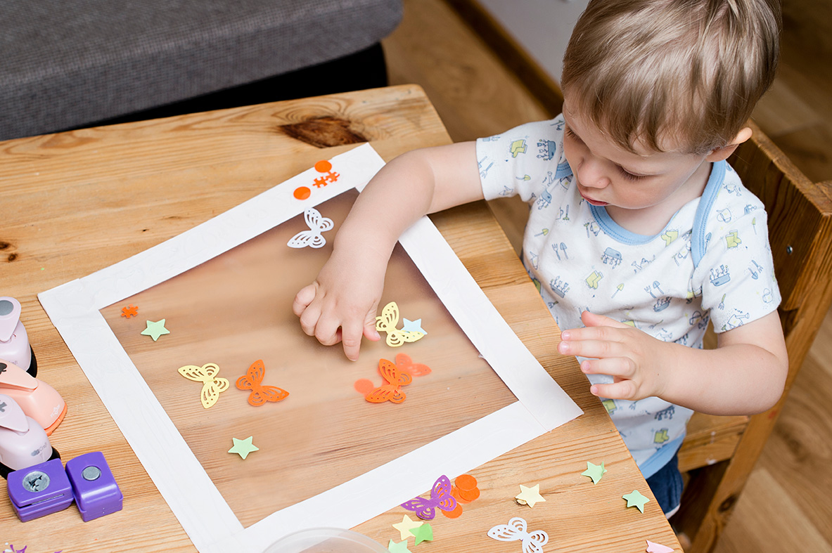 Ein kleines Kind sitzt an einem Holztisch und bastelt mit bunten Papierformen. Es klebt Schmetterlinge, Sterne und andere Formen auf eine transparente Folie, die von einem weißen Rahmen umgeben ist. Auf dem Tisch liegen verschiedene Bastelmaterialien und Locher in Form von Sternen und anderen Motiven. Das Kind ist konzentriert bei der Arbeit und trägt ein hellblau gemustertes T-Shirt.