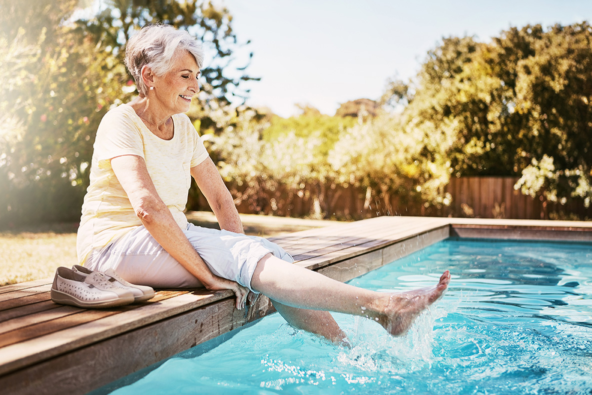 Lächelnde ältere Frau mit grauen Haaren sitzt am Rand eines Pools, trägt ein gelbes T-Shirt und helle Hosen und taucht ihre Füße ins Wasser. Neben ihr stehen ihre Schuhe, während sie die Sonne in einem grünen Garten genießt.