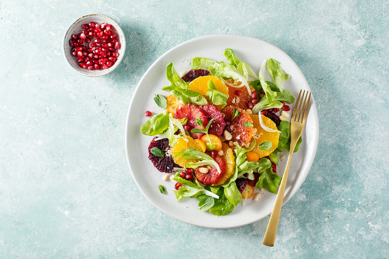 Frischer Zitrussalat mit Blattsalat, Blutorangen-, Orangen- und Mandarinenstücken, Granatapfelkernen, Nüssen und Minzblättern auf einem weißen Teller mit goldener Gabel. Daneben eine kleine Schale mit Granatapfelkernen auf mintgrünem Hintergrund.