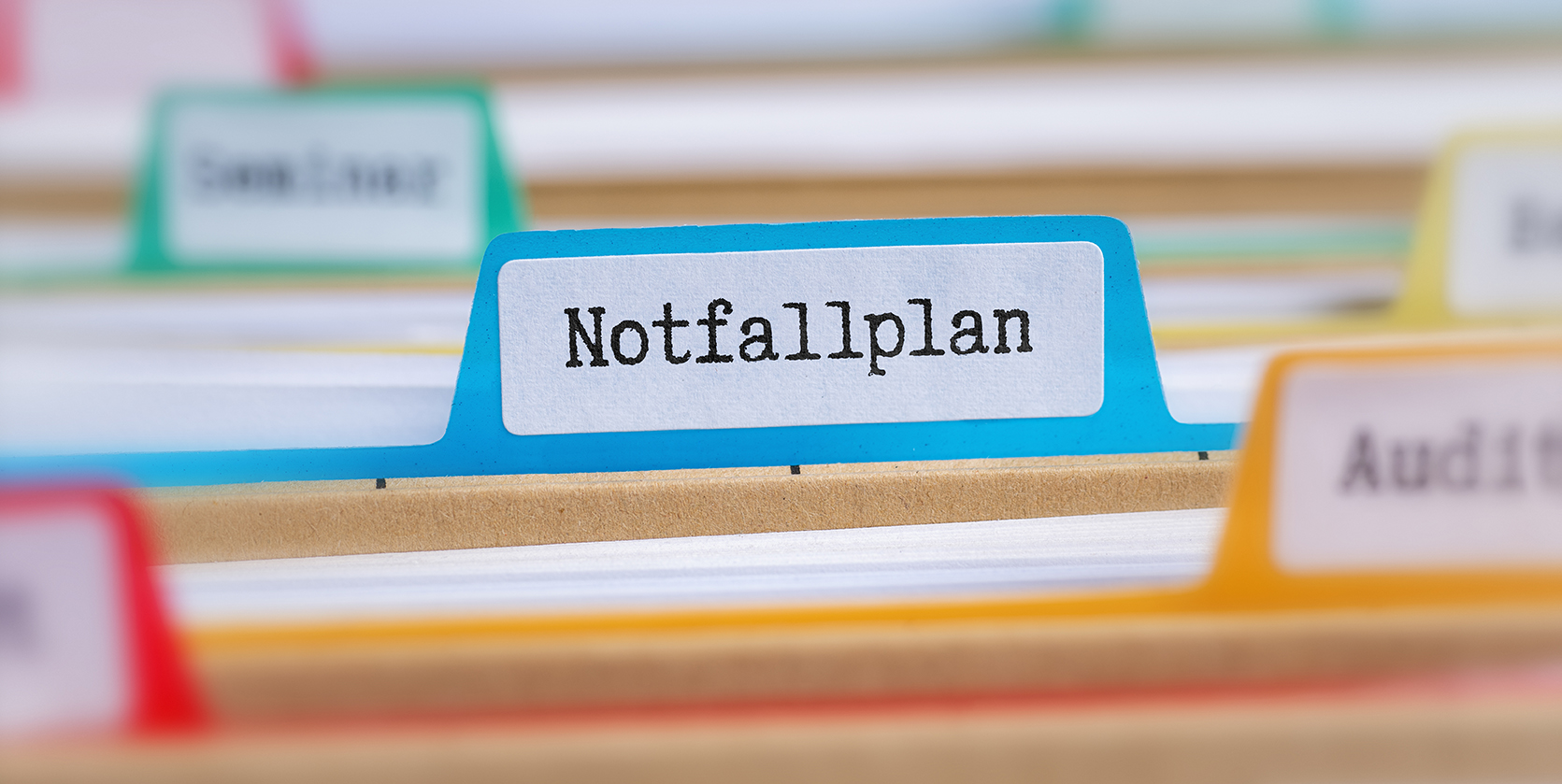 Nahaufnahme eines blauen Registerreiters mit der Aufschrift ‚Notfallplan‘ in schwarzer Schrift. Der Reiter befindet sich in einer Kartei mit weiteren farbigen Registern im Hintergrund, die teilweise unscharf zu erkennen sind.