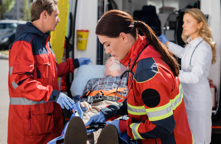 Notfallteam versorgt einen älteren Mann auf einer Trage vor einem Rettungswagen. Zwei Rettungssanitäter in roten Uniformen leisten Erste Hilfe, während eine Ärztin im weißen Kittel im Hintergrund medizinische Geräte vorbereitet. Der Patient trägt ein Sauerstoffgerät, und die Situation wirkt angespannt und fokussiert.