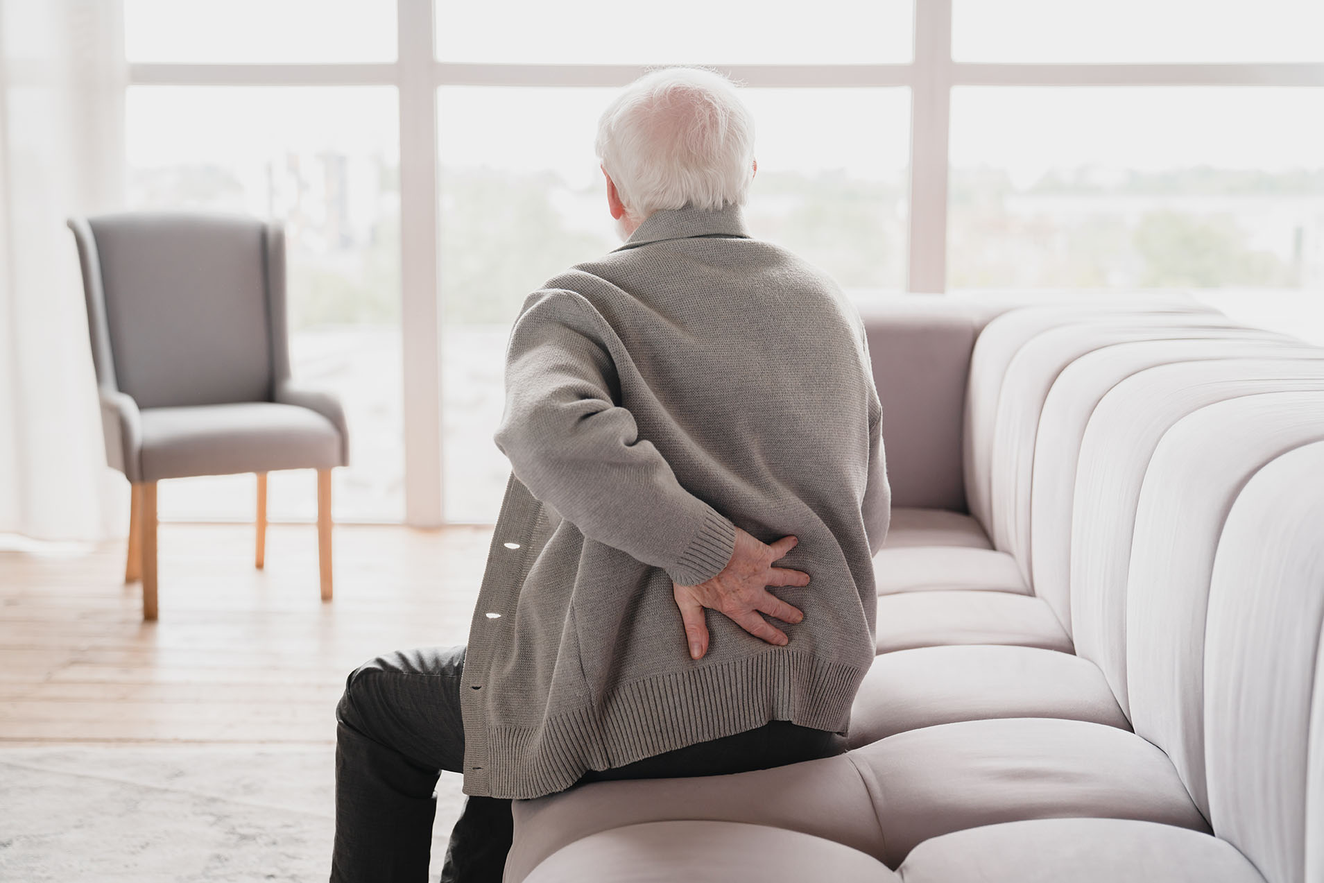 Älterer Mann mit grauen Haaren sitzt auf einem hellen Sofa und hält sich mit schmerzverzerrtem Gesicht den unteren Rücken, vermutlich wegen Rückenschmerzen. Im Hintergrund ist ein großes Fenster mit hellem Tageslicht und ein leerer Stuhl zu sehen.