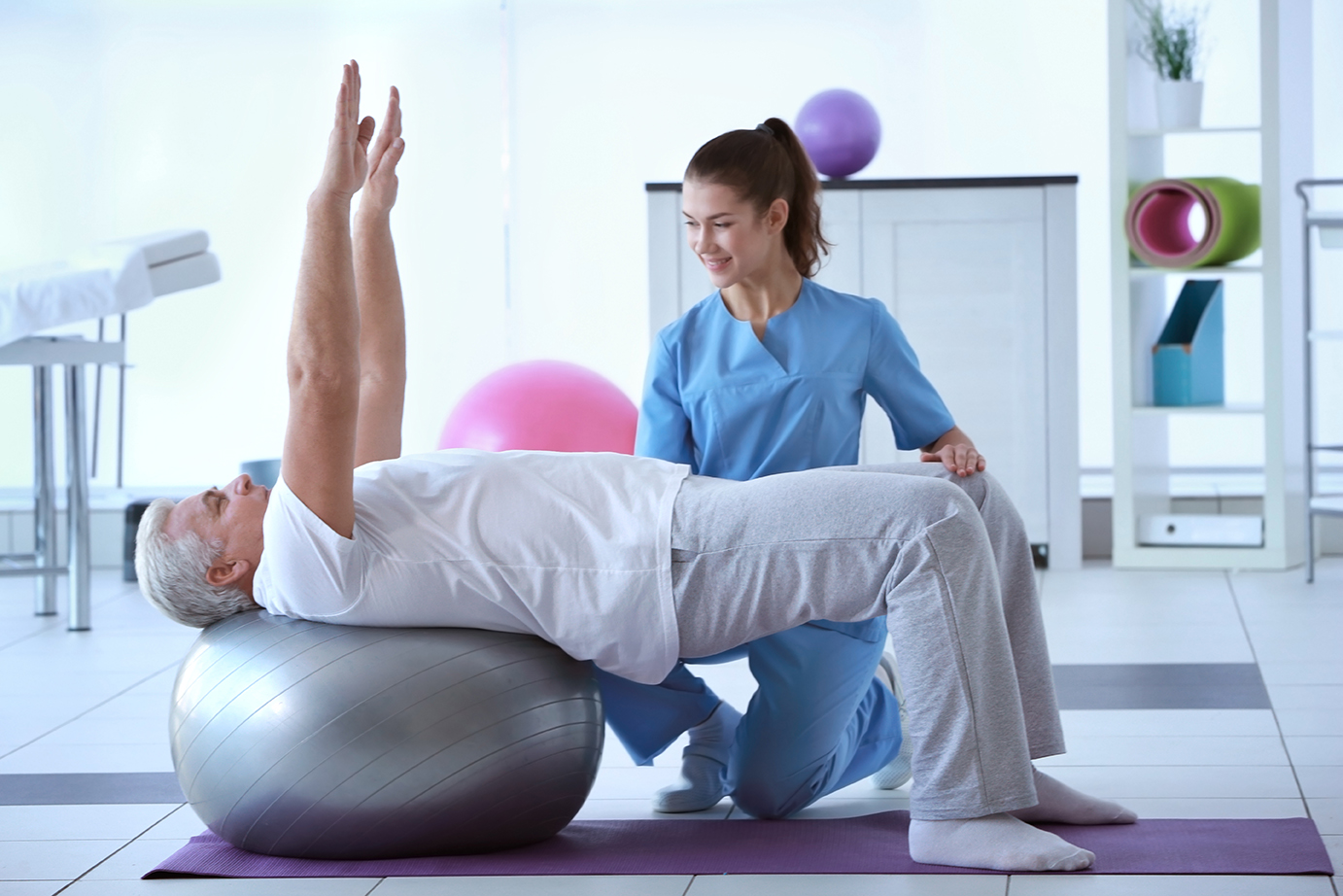 Ein älterer Mann macht unter Anleitung einer Physiotherapeutin Übungen auf einem Gymnastikball. Er liegt mit dem Rücken auf dem Ball, hebt die Arme nach oben und stabilisiert seinen Körper. Die Therapeutin kniet daneben und gibt Anweisungen. Die Szene spielt sich in einem modernen Physiotherapie-Raum mit Trainingsgeräten und Gymnastikbällen im Hintergrund ab.
