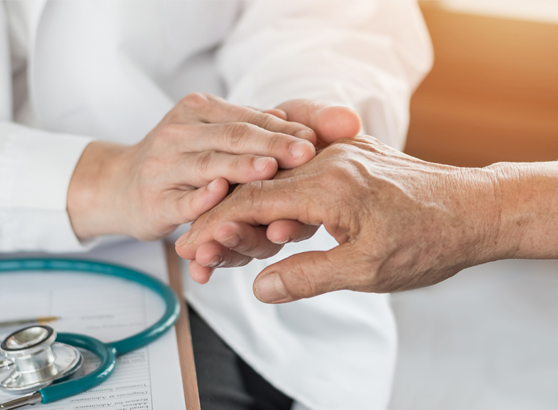 Eine Ärztin oder Pflegekraft hält einfühlsam die Hand eines älteren Patienten als Zeichen von Fürsorge und Unterstützung. Im Hintergrund liegt ein Stethoskop auf Dokumenten.