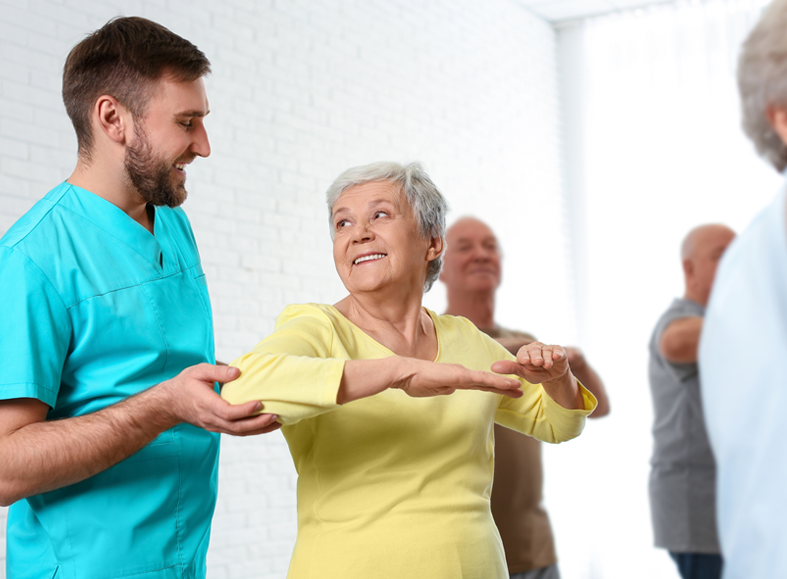 Eine ältere Frau in einem gelben Oberteil nimmt mit Freude an einer Bewegungsübung teil, während ein freundlicher Physiotherapeut oder Pflegekraft sie unterstützt. Im Hintergrund machen weitere Senioren mit.