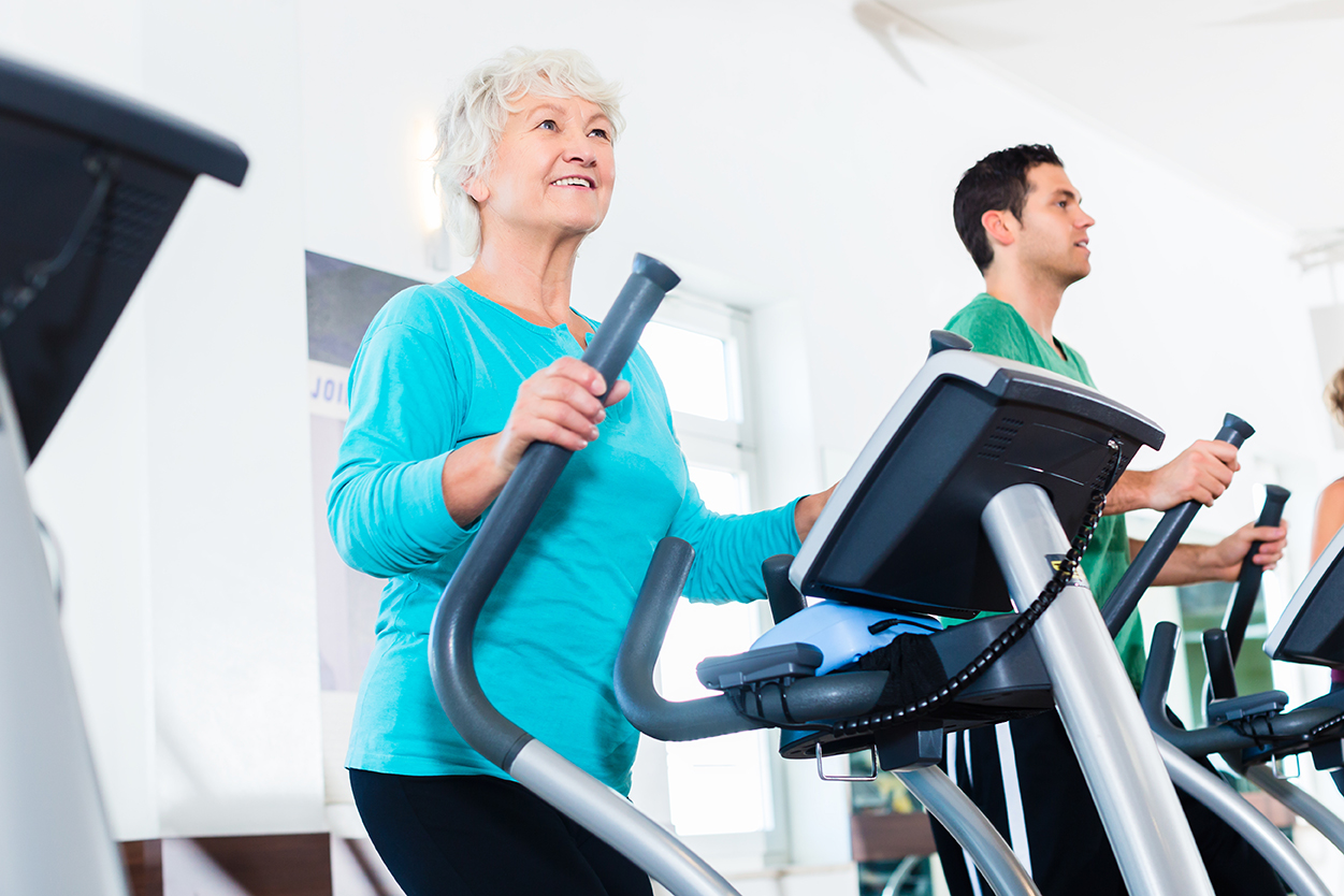 Ältere Frau trainiert motiviert auf einem Crosstrainer im Fitnessstudio, während ein jüngerer Mann neben ihr ebenfalls trainiert. Symbol für Aktivität, Gesundheit und körperliche Fitness im Alter.