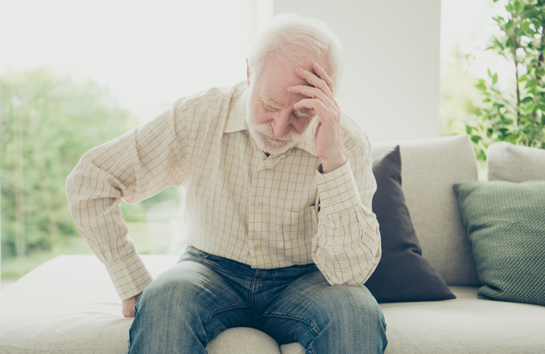 Älterer Mann mit weißem Haar und Bart sitzt auf einem Sofa. Er trägt ein kariertes Hemd und Jeans, hält sich mit einer Hand den unteren Rücken und stützt den Kopf mit der anderen Hand, was auf Schmerzen oder Erschöpfung hindeutet. Im Hintergrund sind Kissen und ein Fenster mit Blick ins Grüne zu sehen.